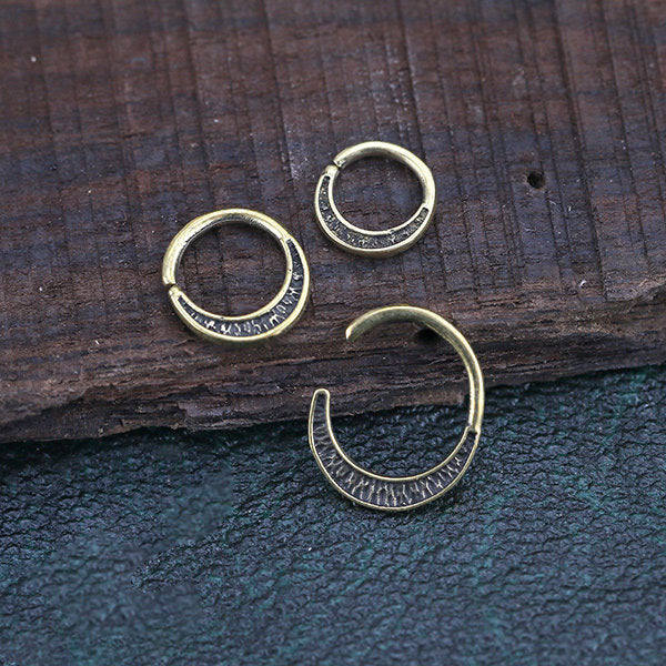 Three small MOON Seamless Rings in gold, made by Kaya Movement, with a hammered texture design lay on a wooden and leather surface. Two rings are closed, one is open, showcasing detailed metalwork. These minimalist gold brass body jewelry rings have a simple yet elegant look with an antique aesthetic.
